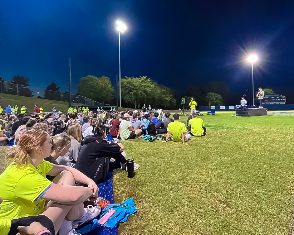 Fields of Faith Event. Students of all ages are sitting on the Crowder Field grass as they listen to Mikey Brinton speak.