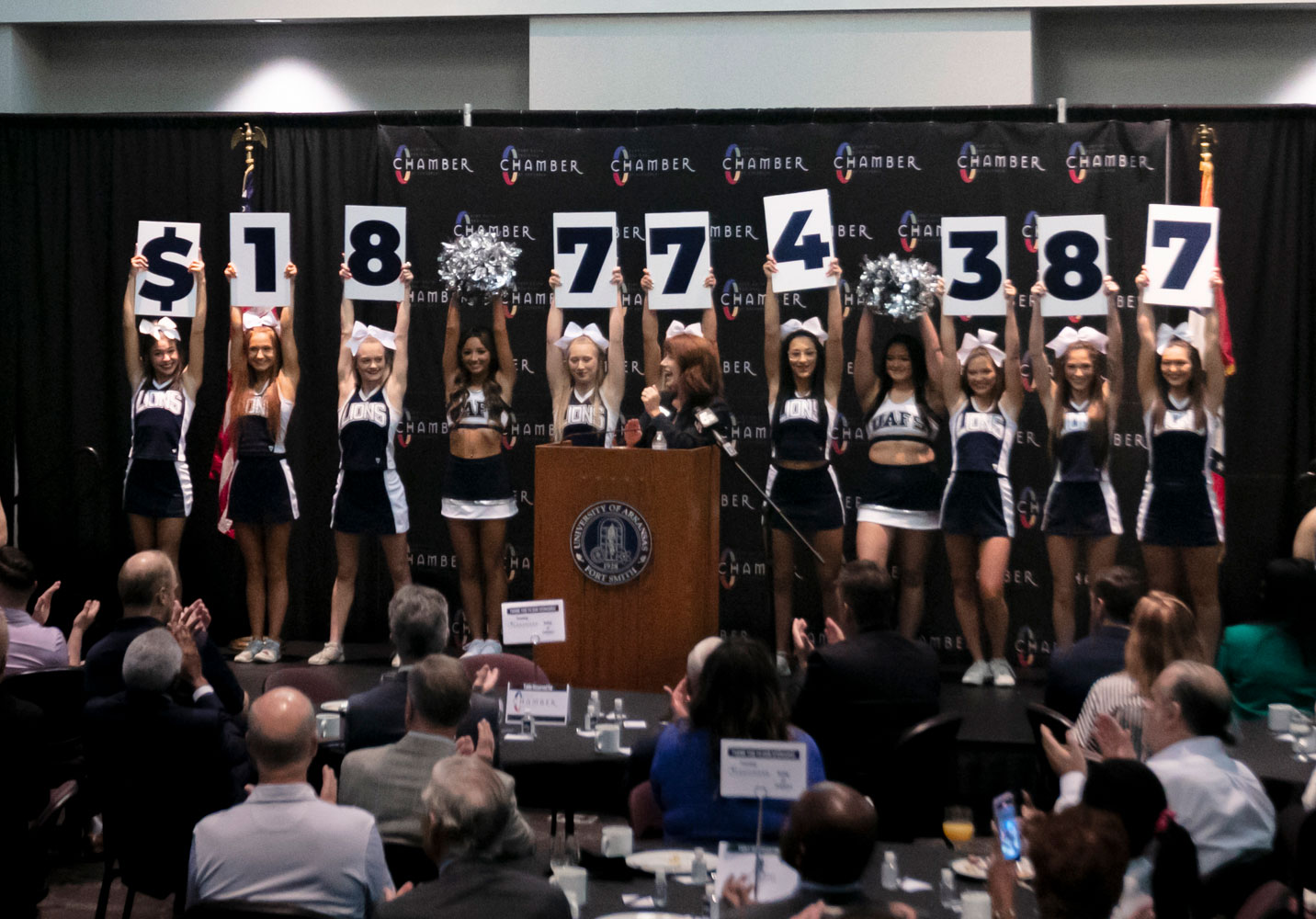 Eleven cheerleaders hold signs that read $18,774,387 on stage at gift announcement. 